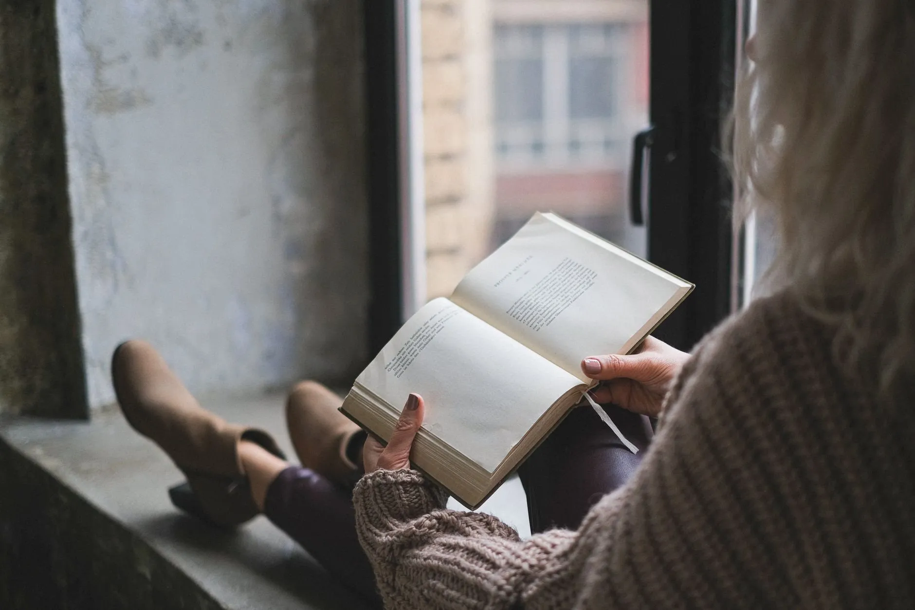 A reader sitting on a deep windowsill with an open book on her knees, seen from behind